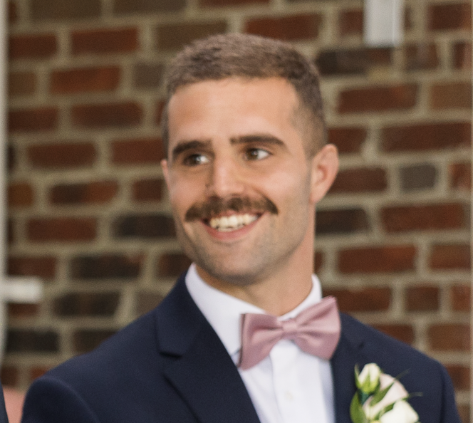 A man with a mustache smiling, dressed in a navy blazer, white shirt, and pink bow tie, standing in front of a brick wall.