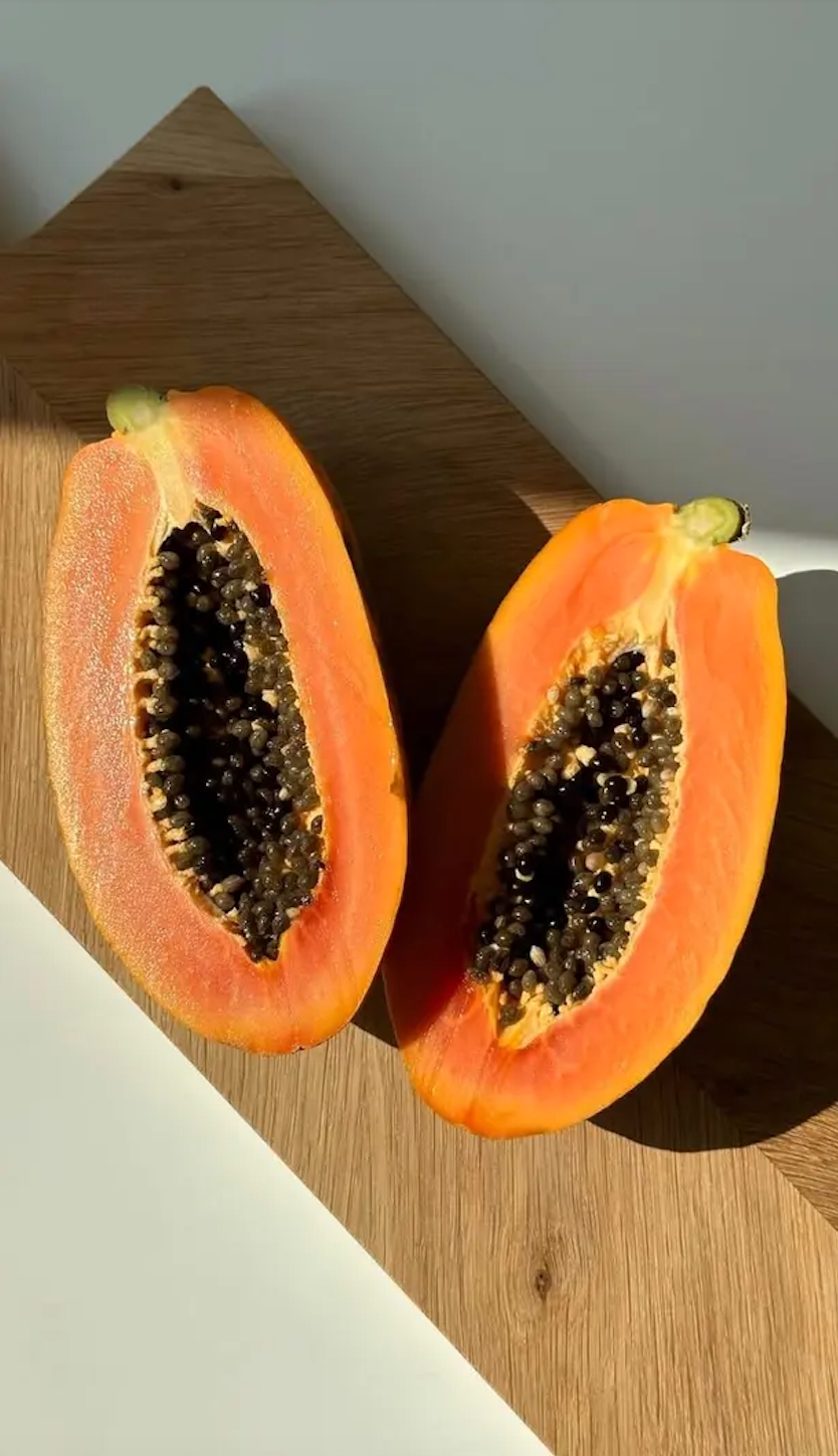 Half of a ripe papaya fruit with orange flesh and black seeds on a wooden cutting board, with sunlight casting shadows.
