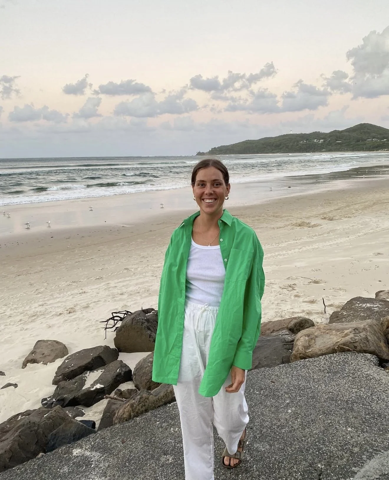 A woman smiling at the camera on a beach, wearing a bright green shirt over a white tank top and white pants, with rocks on the shore and the ocean in the background.