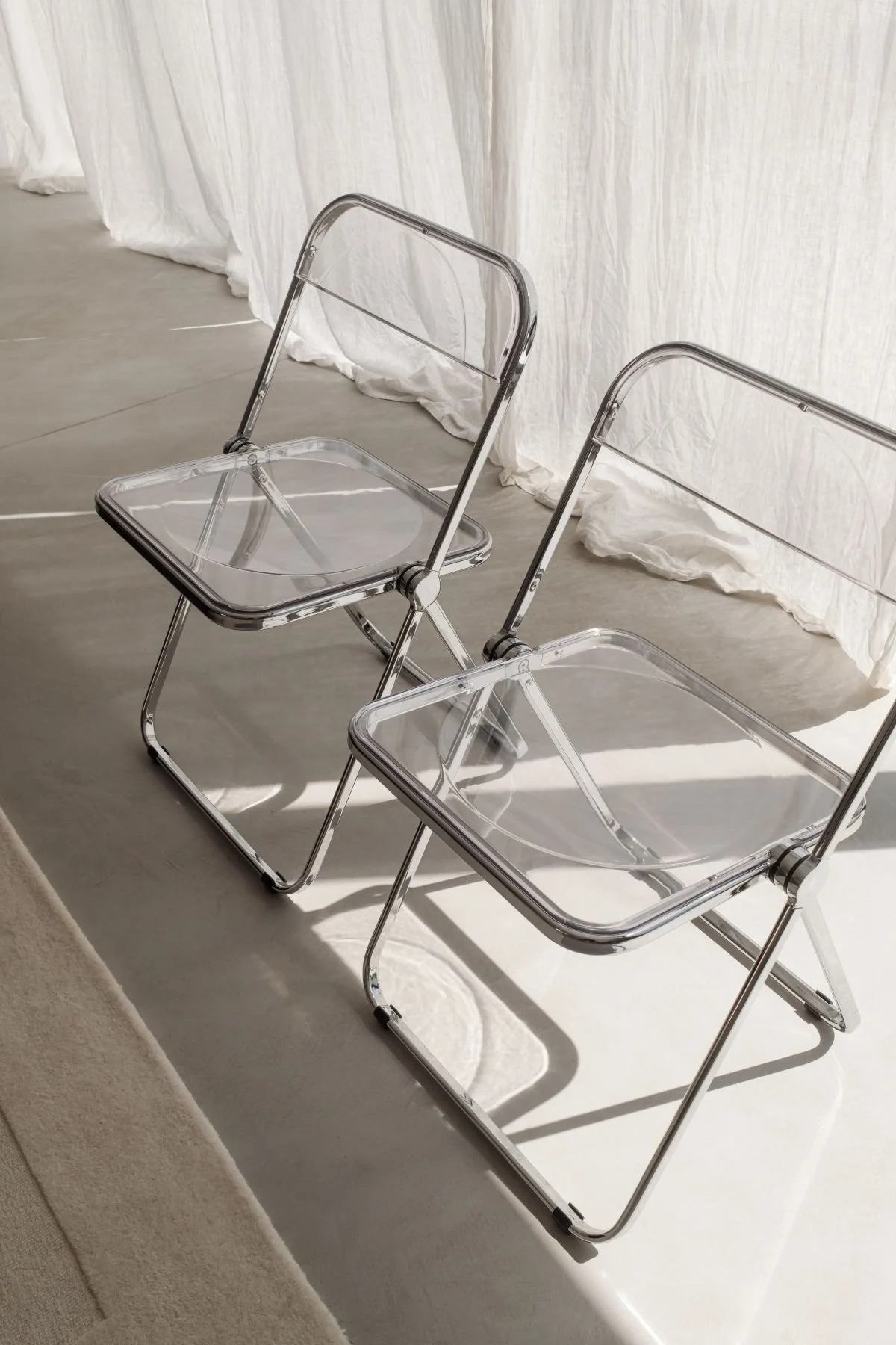 Two clear acrylic folding chairs with metal frames on a light-colored floor near a white curtain.