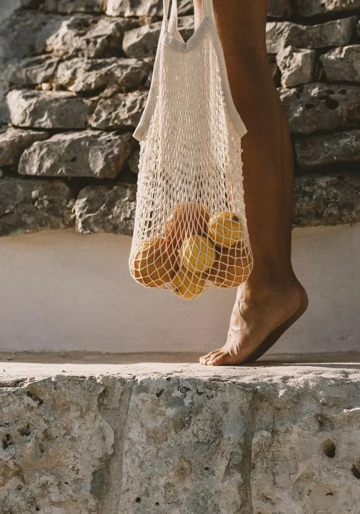 A person walking barefoot on a stone surface, carrying a mesh bag filled with yellow lemons, against a background of a stone wall.