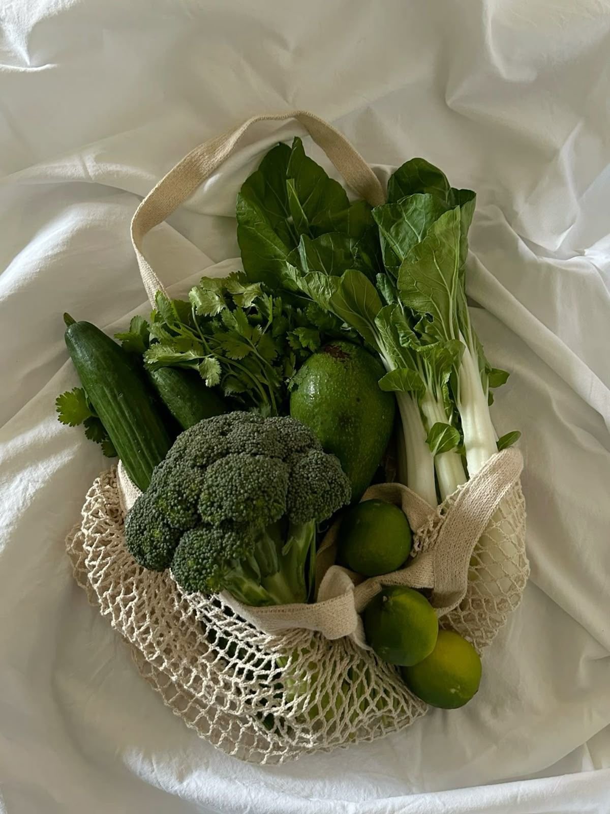 A reusable shopping bag filled with fresh green vegetables and fruits, including broccoli, cucumbers, cilantro, lettuce, limes, and green onions, resting on a white fabric surface.