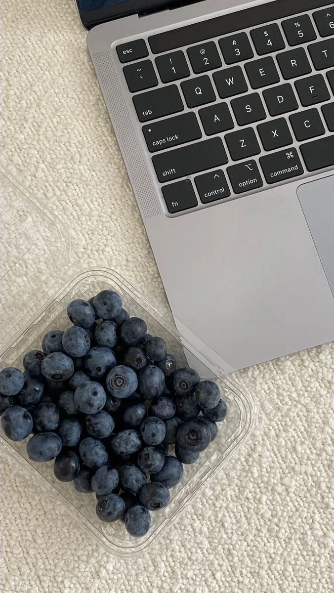 A close-up of a silver laptop keyboard next to a plastic container of blueberries on a textured beige surface.