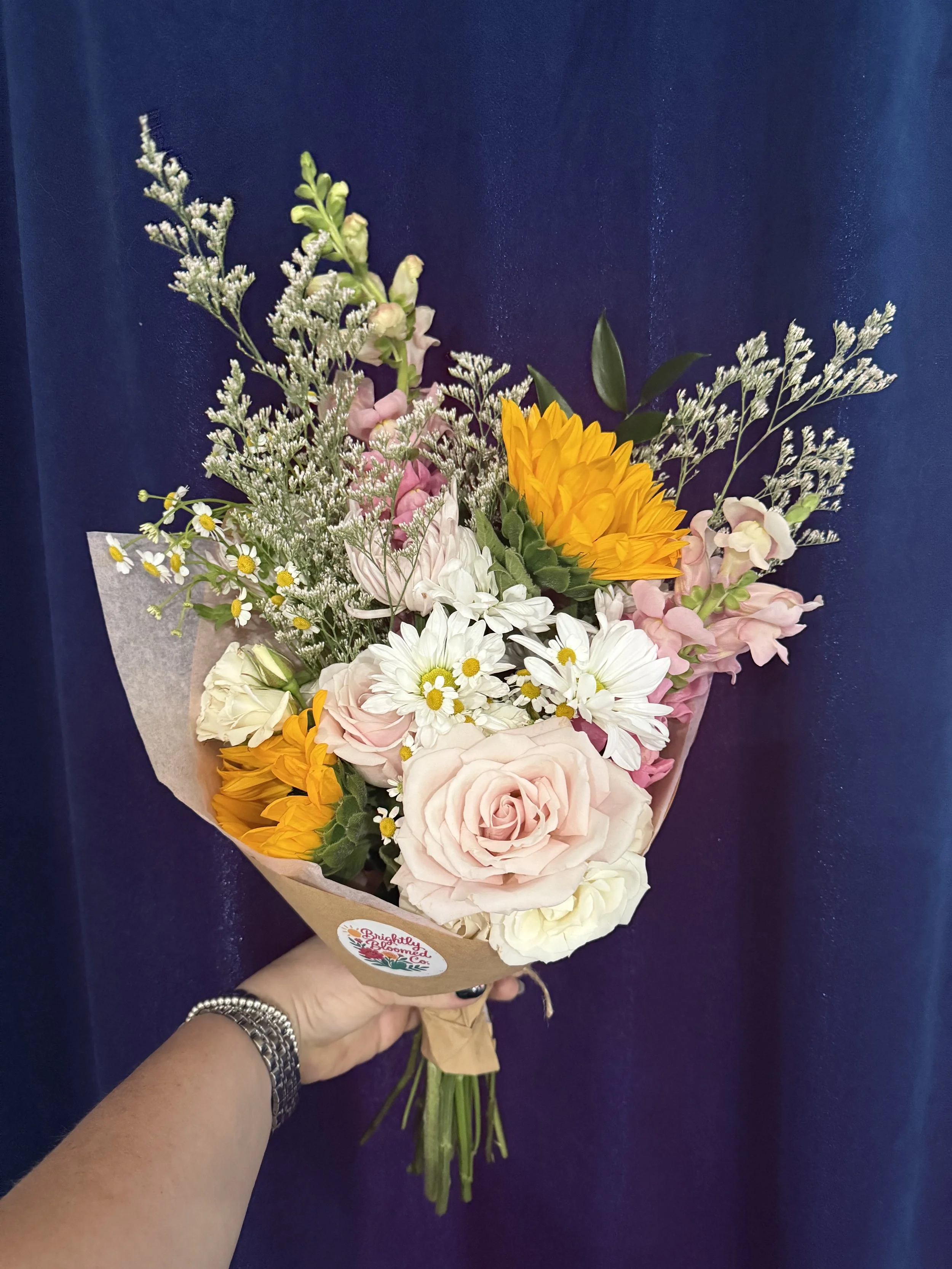 A hand holding a colorful bouquet of flowers against a dark blue background. The bouquet includes pink roses, white daisies, yellow sunflowers, and filler greenery. All Arrangements are custom and made to order. Delivery is available.