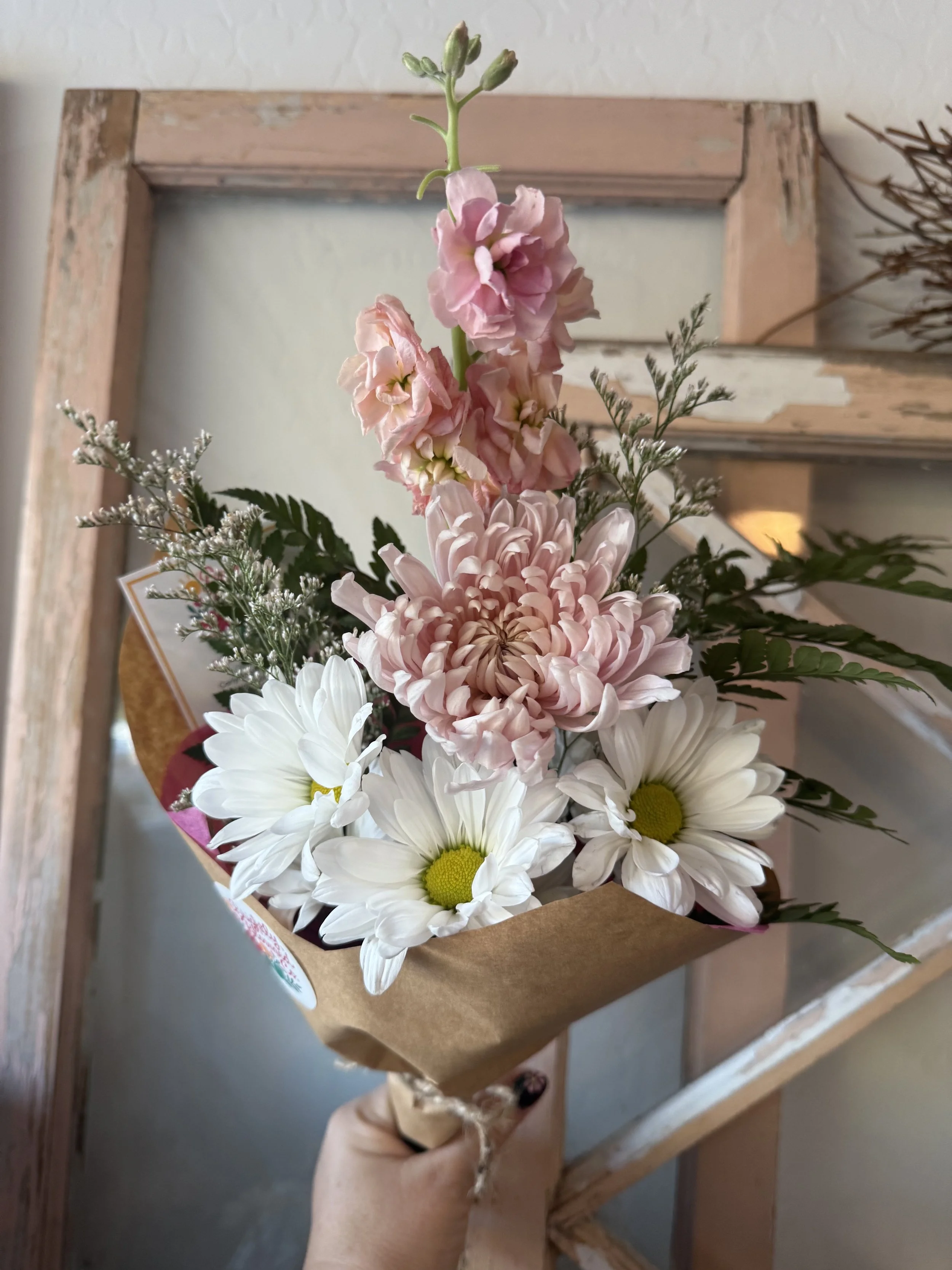 Hand holding a bouquet of pink, white, and green flowers against a wooden background. All Arrangements are custom and made to order. Delivery is available.