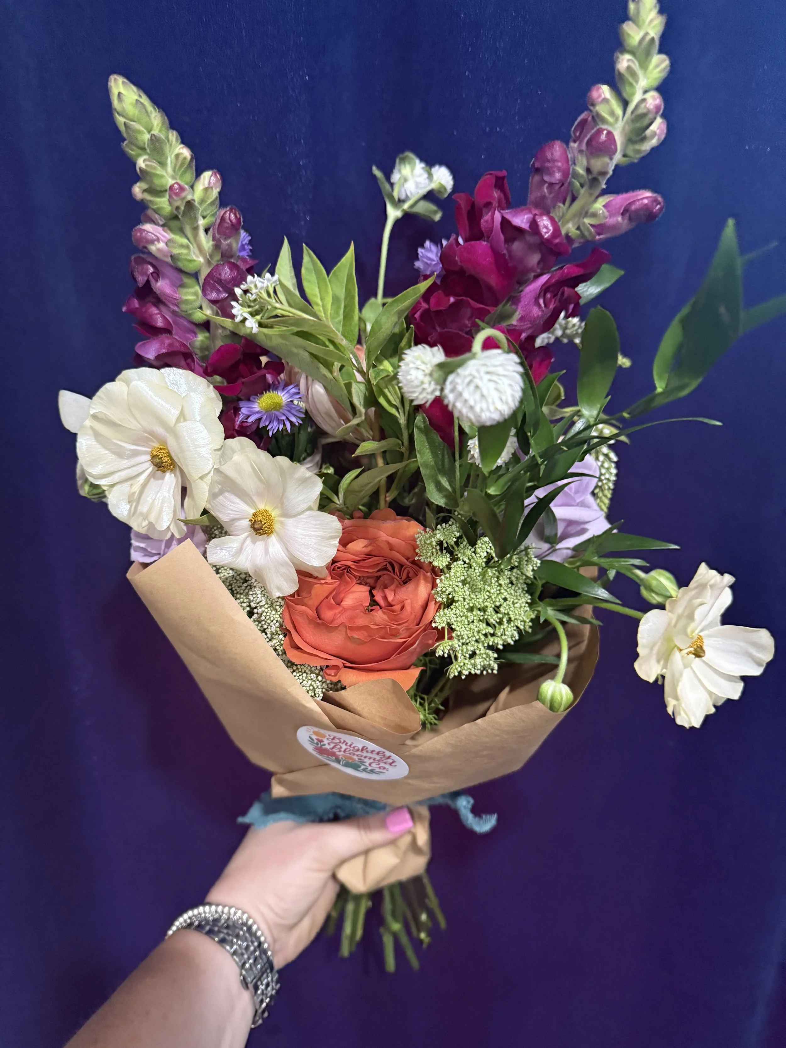 A hand holding a bouquet wrapped in brown paper against a dark blue background. The bouquet contains white, purple, pink, and orange flowers, with various green leaves and foliage. All Arrangements are custom and made to order. Delivery is available.