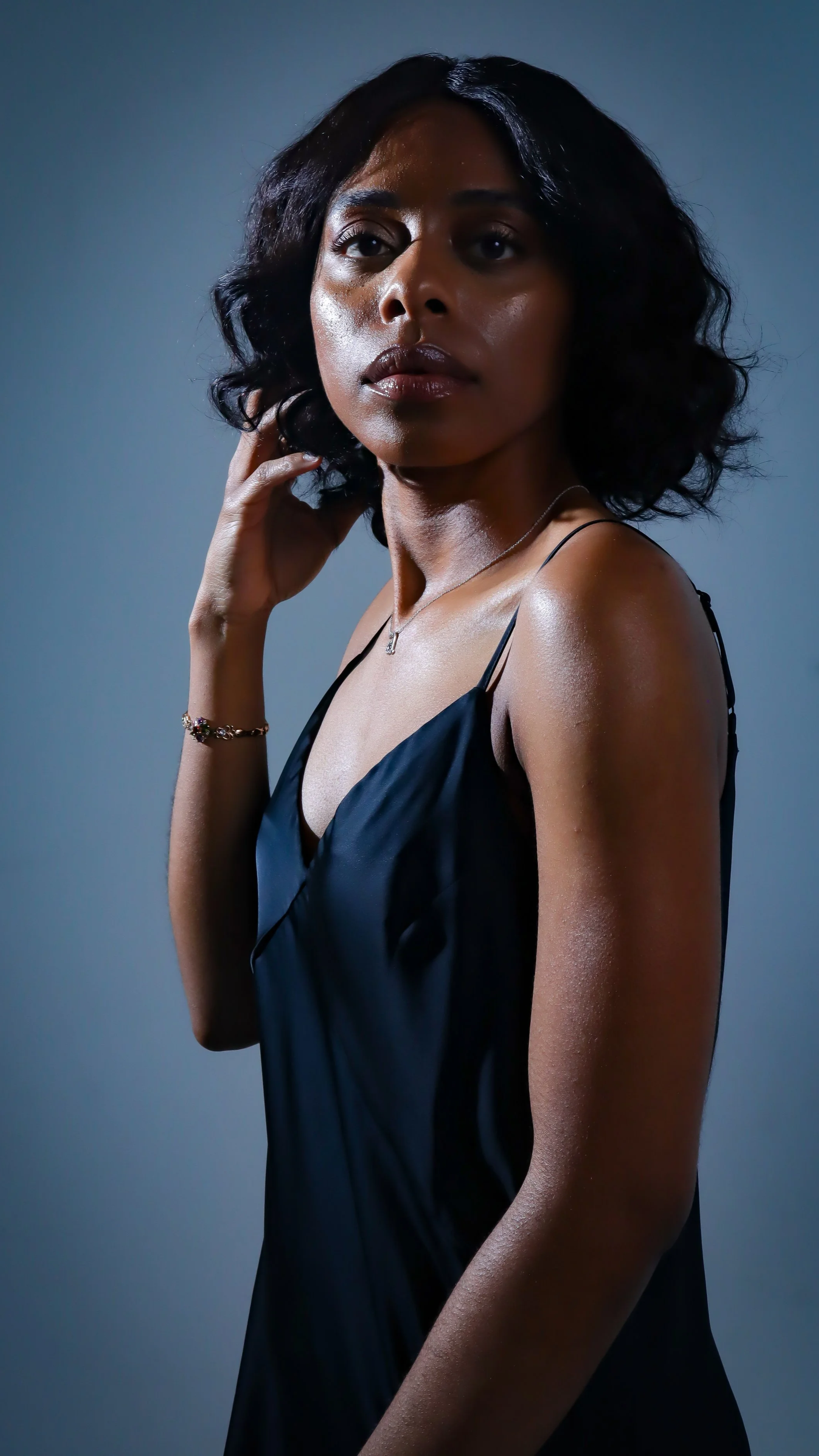 A woman with black, wavy hair wearing a black silky spaghetti strap dress, with jewelry including a necklace and bracelet, posing against a plain background.