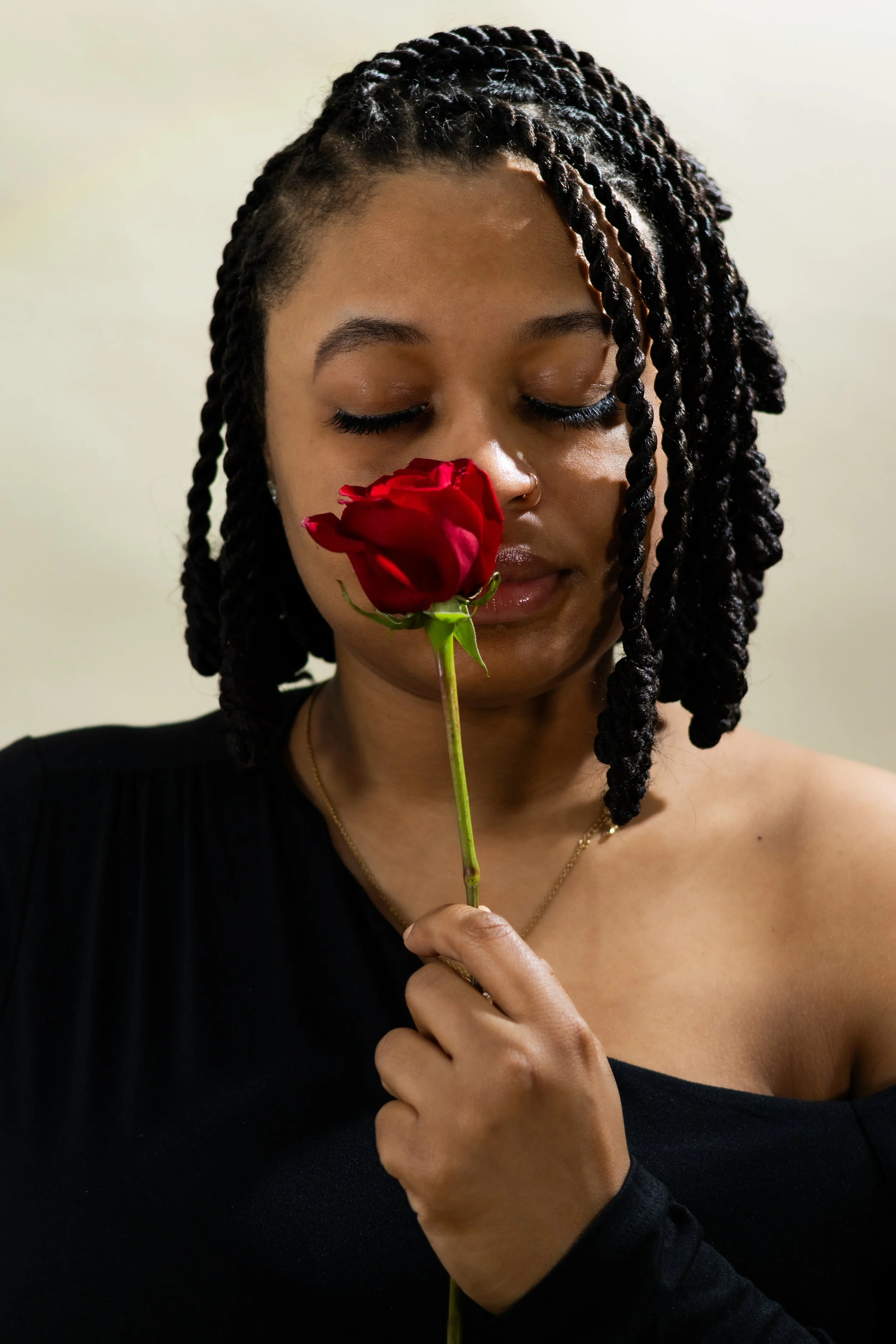 A woman with styled black hair holds a red rose near her face with her eyes closed.
