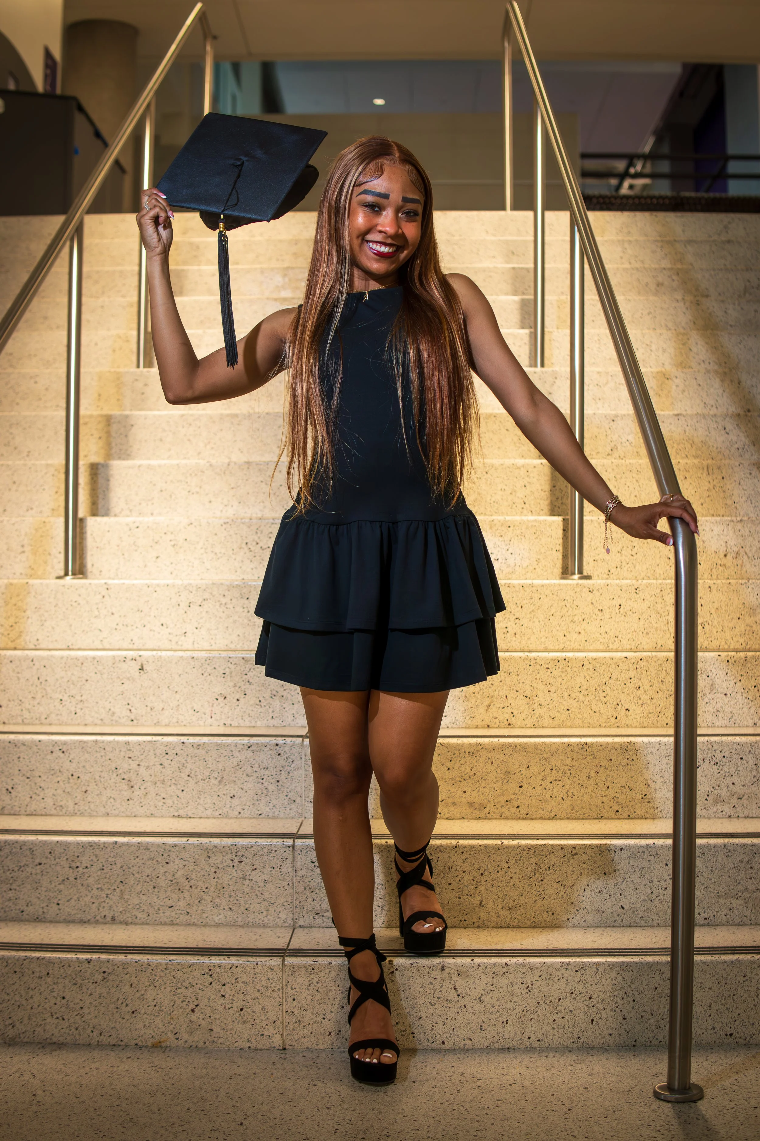 A young woman in a sleeveless black dress and black high-heeled shoes is standing on a staircase, smiling and holding a graduation cap in one hand. She appears to be celebrating her graduation.