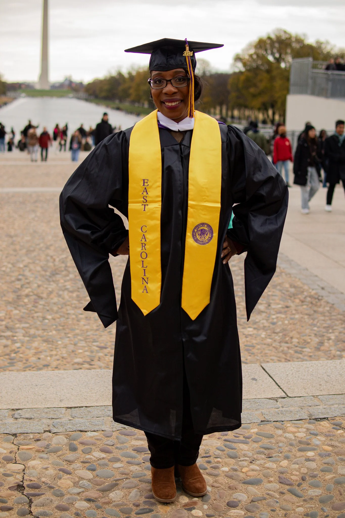 Woman in graduation gown and cap with a yellow stole that says 'East Carolina' and a university emblem, standing outdoors during the day with trees and other people in the background.
