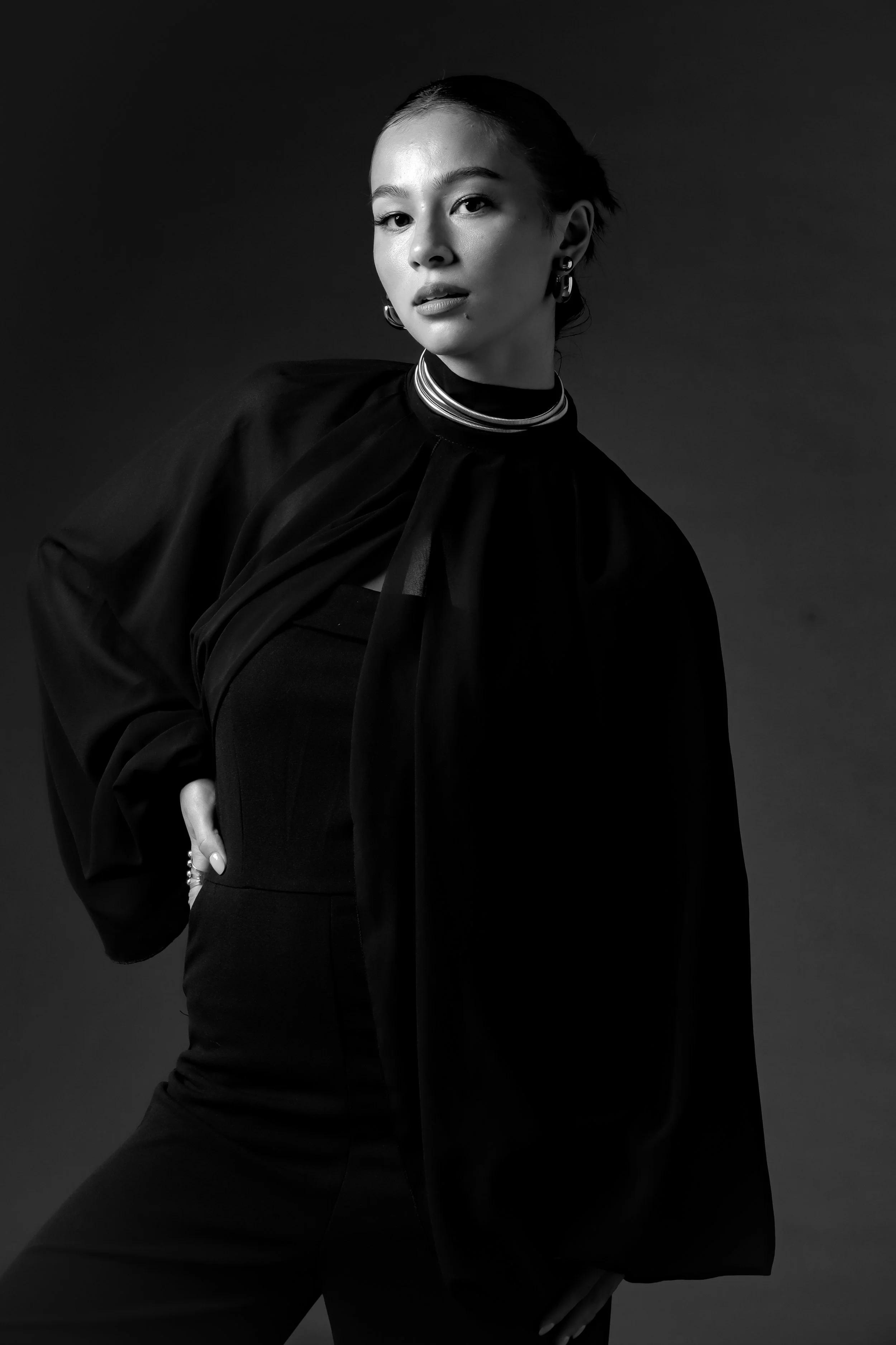 Black and white portrait of a woman wearing a high-neck top, a flowing jacket, and jewelry, looking confidently at the camera.