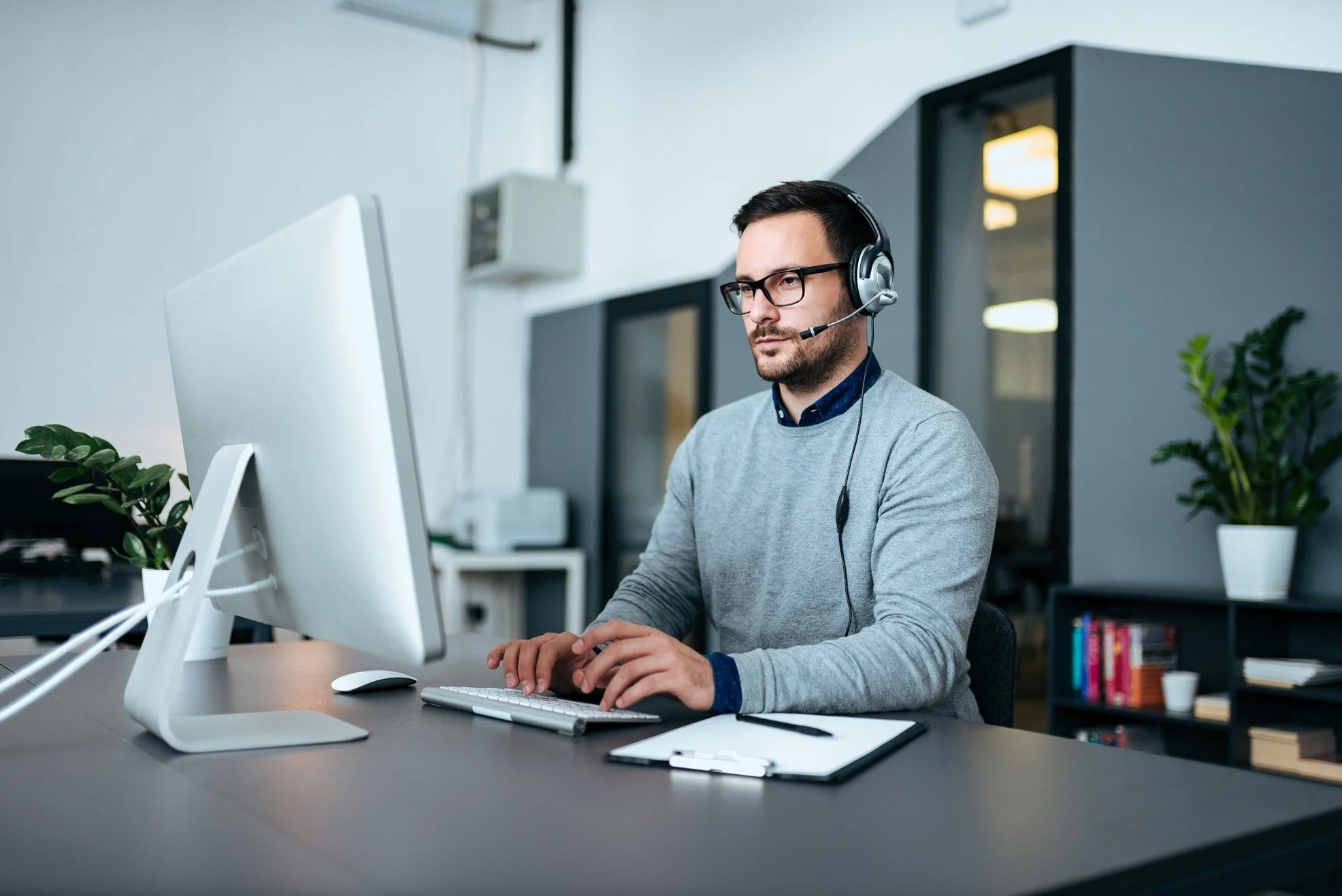 Man sitting at a desk working on a computer with a headset on, in a modern office setting.