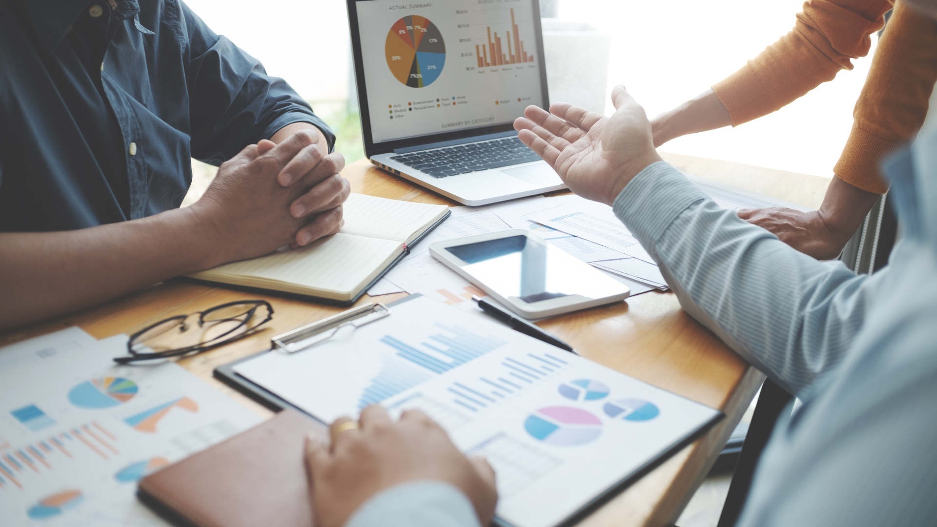 A business meeting with multiple people discussing at a desk with charts, graphs, a laptop, tablet, and notebook.