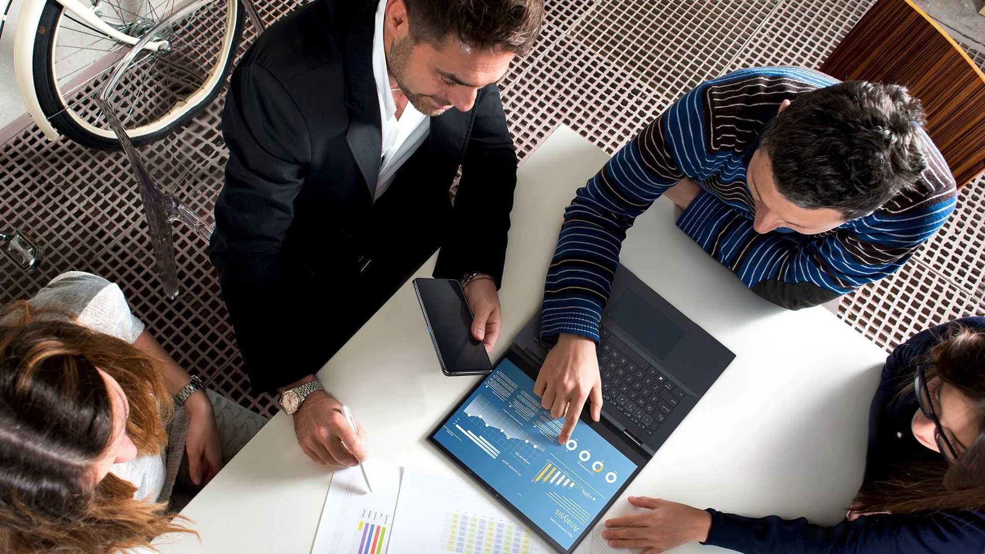 A group of five professionals in a business meeting gathered around a table, looking at a laptop displaying charts and graphs with data analysis.