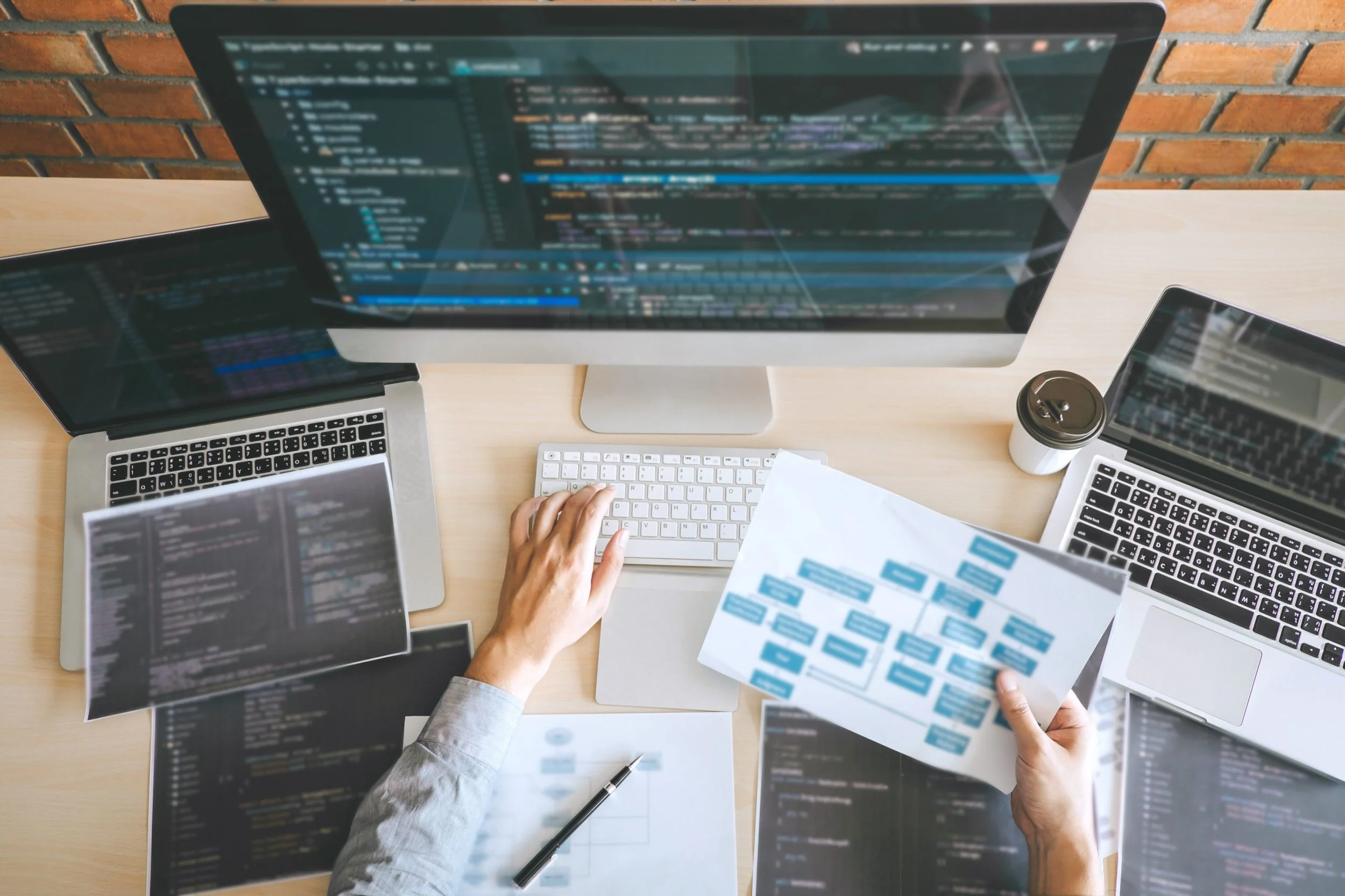 Multiple computer screens displaying lines of code and software development tools, with a person's hand holding a printed flowchart or diagram, and a laptop and a coffee cup on a light wooden desk against a brick wall background.
