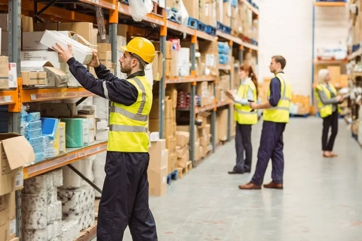Workers in safety vests and helmets stocking shelves in a warehouse store aisle with boxes and products.