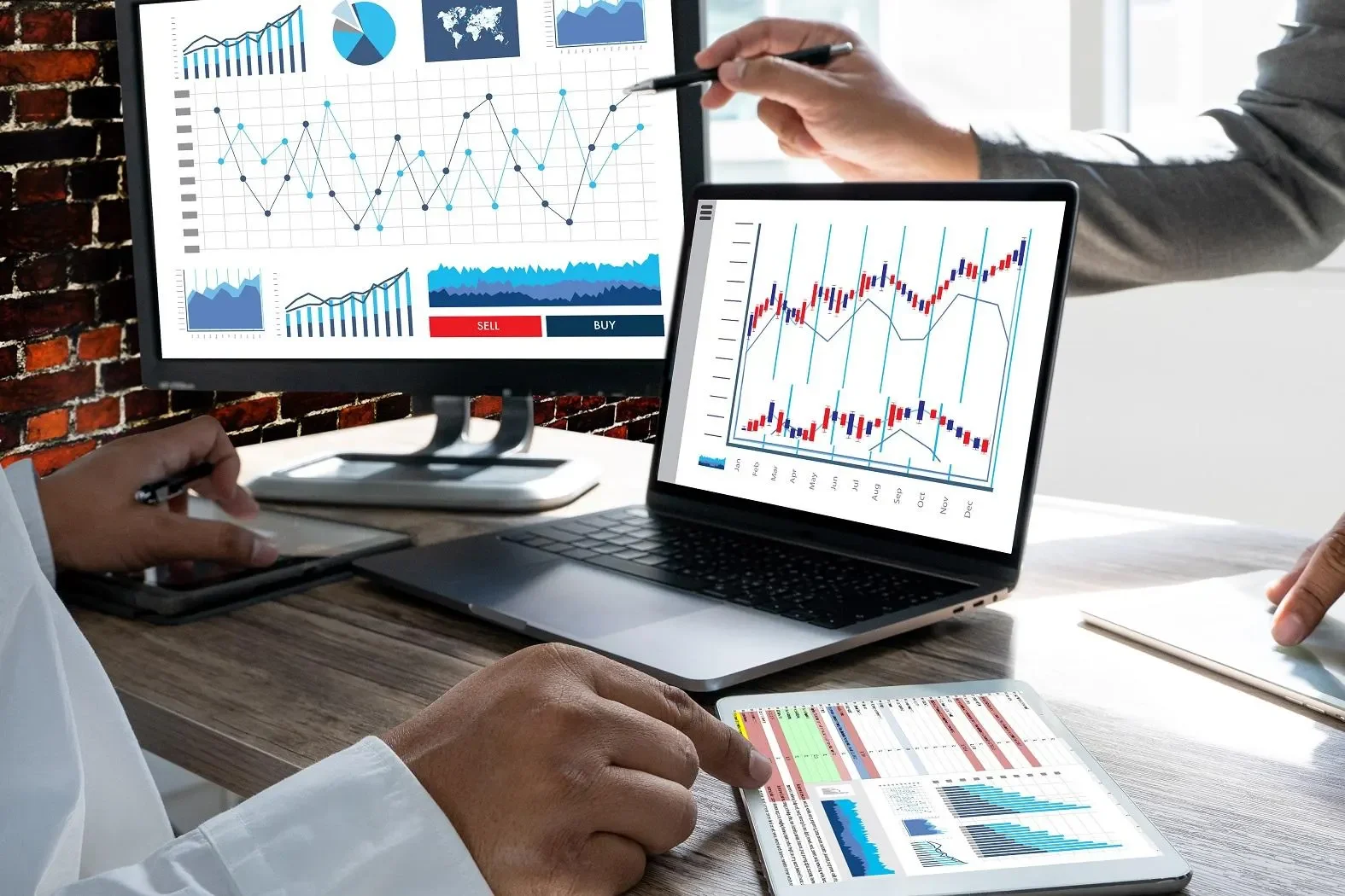 Business meeting with multiple digital devices displaying financial charts and data, including line graphs, bar charts, and pie charts, on a wooden table against a brick wall background.