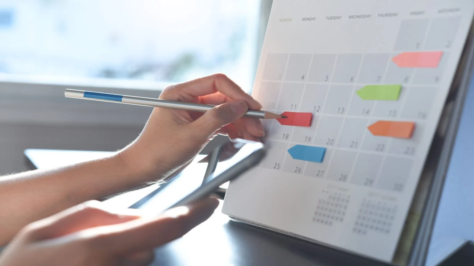 A person holding a smartphone and a pen, marking a red sticker on a desk calendar with a laptop in the background.