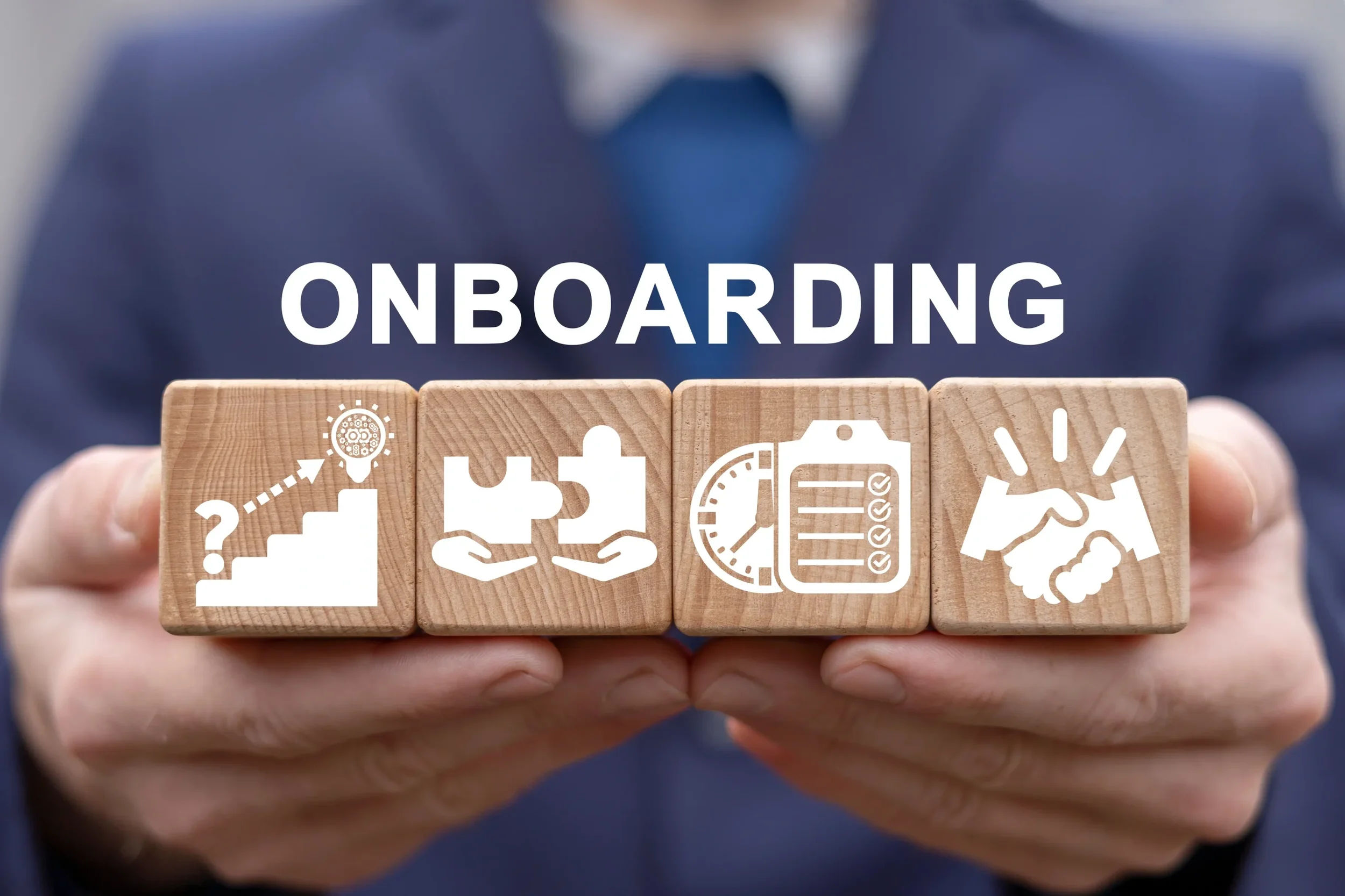 Close-up of a hand holding five wooden blocks with white icons and the word 'ONBOARDING' on top. The icons depict a staircase with a question mark, a gear with a dotted arrow, a pair of hands holding a puzzle piece, a clock and checklist, and a handshake.