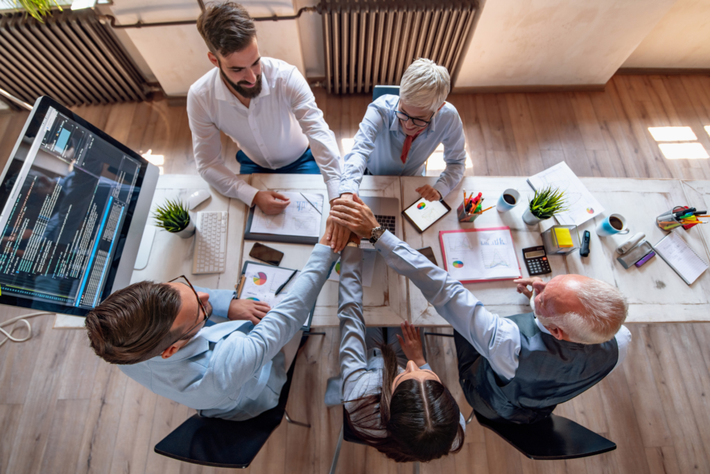 Businesspeople in a meeting placing their hands together over a table with documents, laptops, and office supplies.
