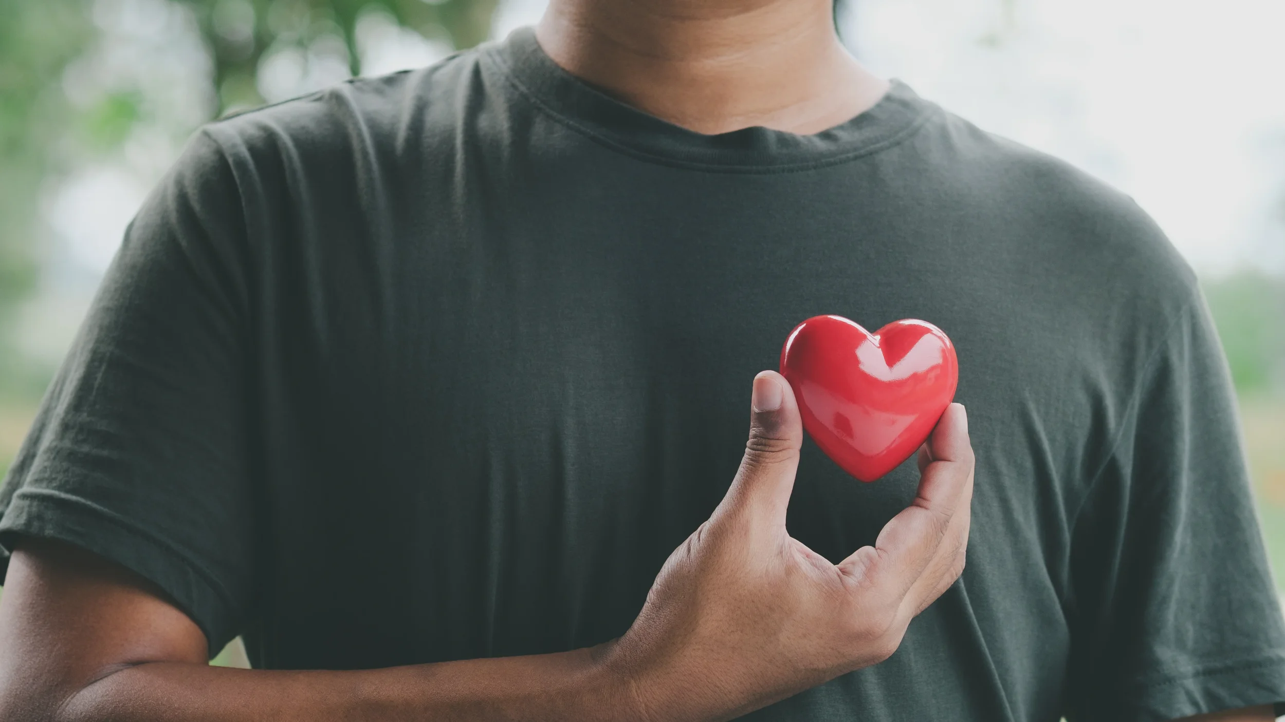 Person wearing a dark shirt holding a red, shiny, ceramic heart with their right hand outside with greenery in the background.