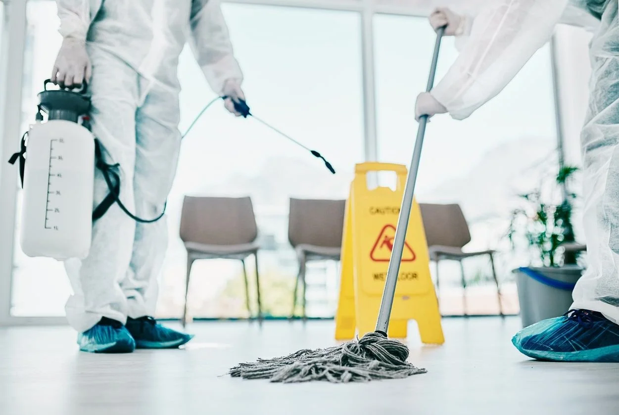 People in protective suits disinfecting a floor with cleaning equipment and a wet mop, while a yellow wet floor sign is nearby.