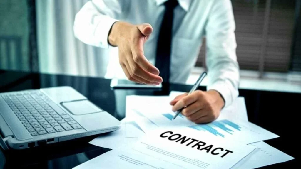 A person in a business shirt and tie is sitting at a desk, signing a contract with a pen, with a laptop and documents on the desk.
