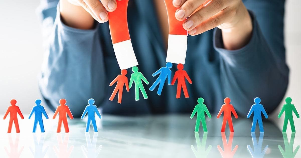 Person holding a magnet attracting paper cutouts of green, red, and blue human figures, with more figures on the table.
