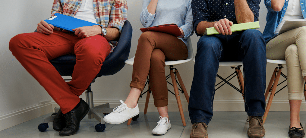 Four people sitting on chairs in a classroom or waiting area, holding notebooks or folders, dressed casually.