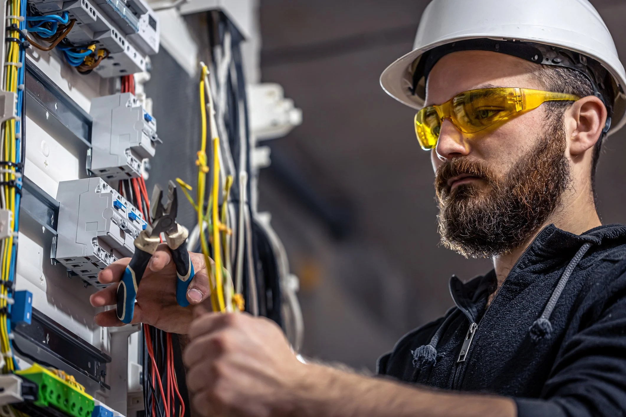 A male electrician with a beard, wearing a white hard hat and yellow safety glasses, working on an electrical panel with multicolored wires and circuit breakers.