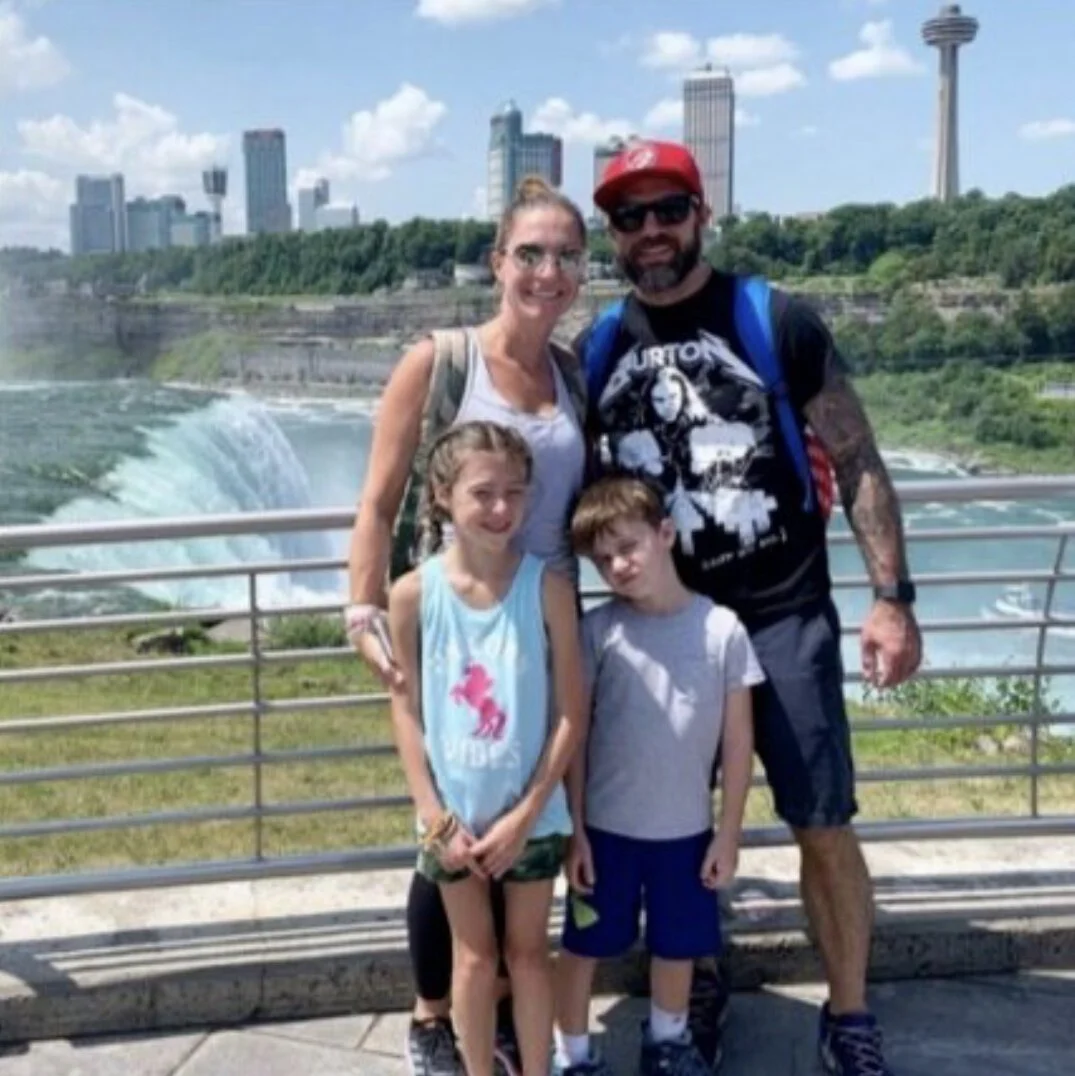 A family of four posing in front of Niagara Falls with the city skyline in the background on a sunny day.