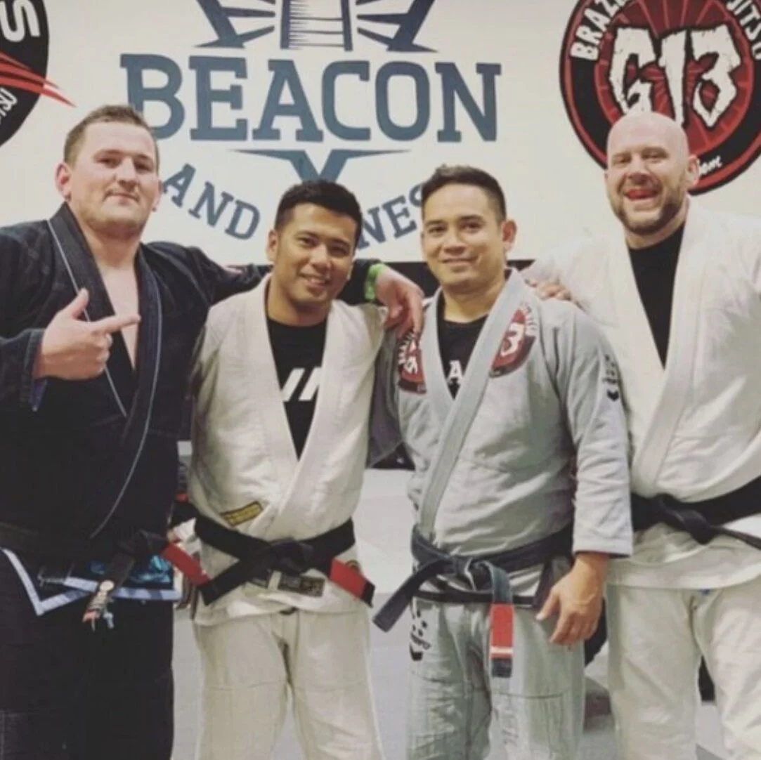 Four men wearing Brazilian Jiu-Jitsu gis and black belts, standing together and smiling in front of a gym sign.