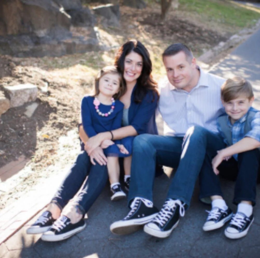 A family of four sitting on a path outdoors, all wearing casual clothing and black Converse shoes. The mother and daughter are on the left, while the father and son are on the right. They appear happy and relaxed.