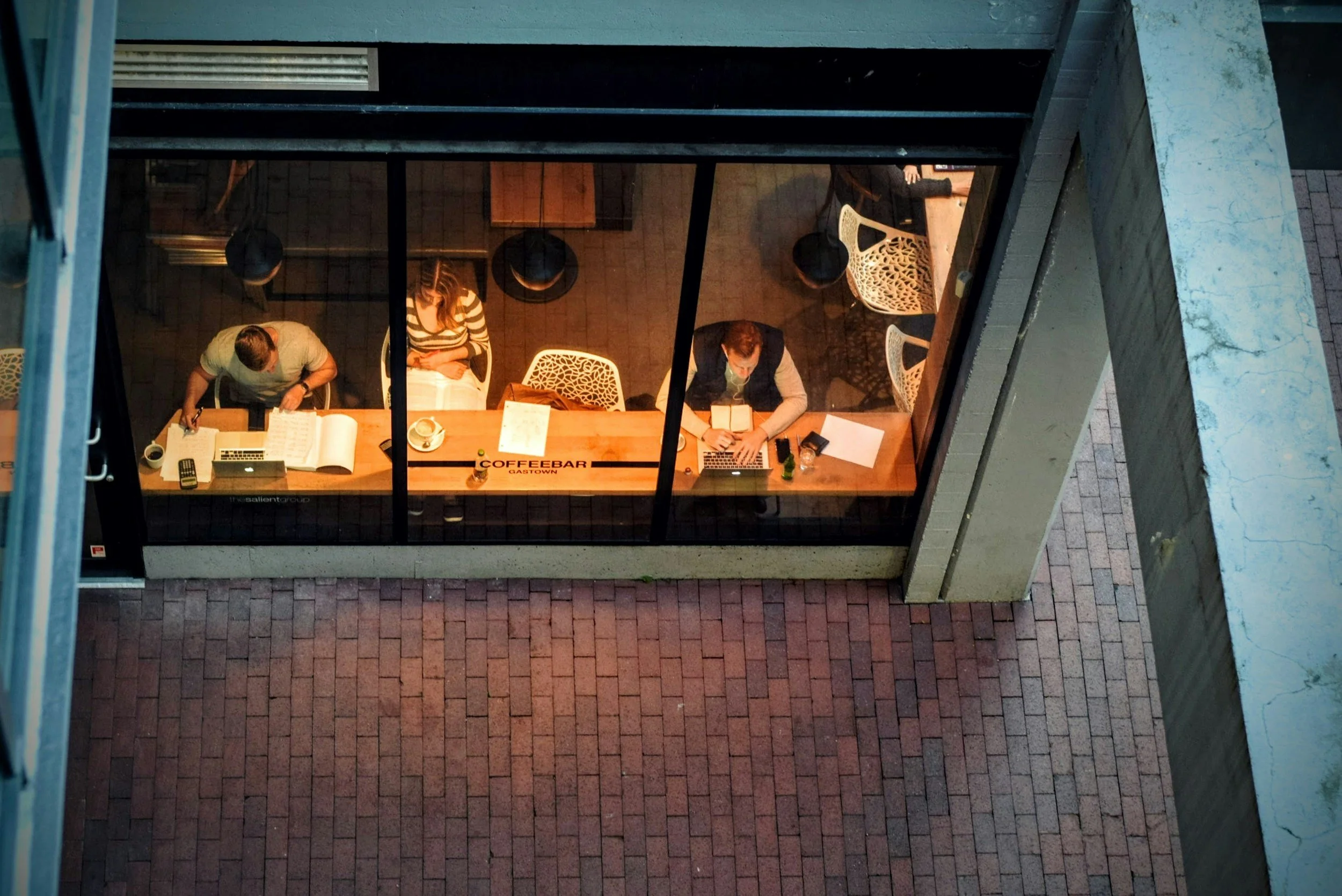 View from above of three people working at a coffee shop table seen through large glass windows, with a brick sidewalk outside.