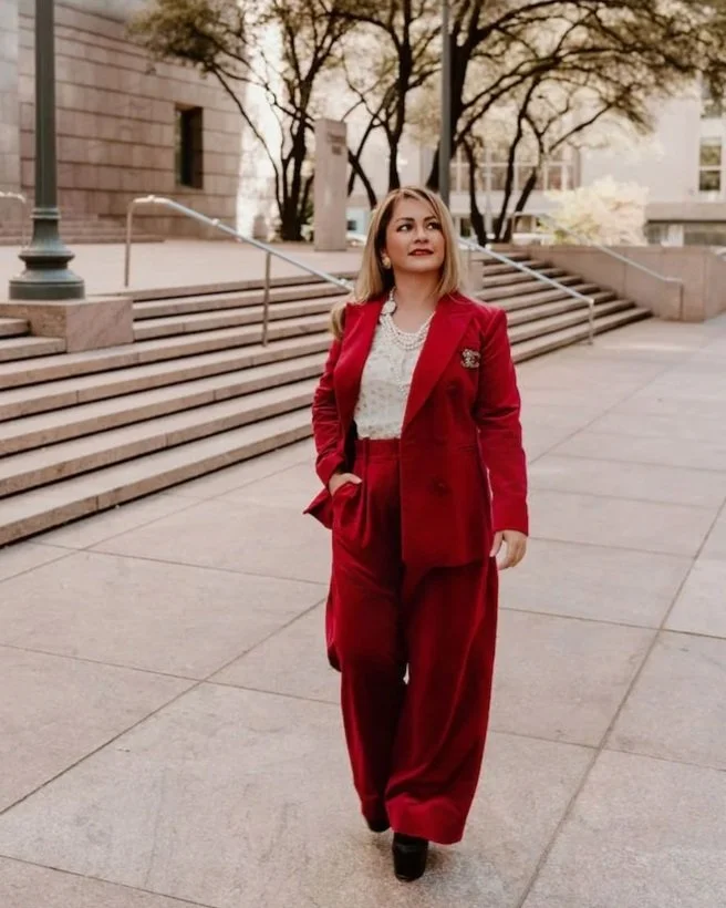 A woman in a bright red pantsuit, white blouse, and pearl necklace walking outside near large stairs and trees.
