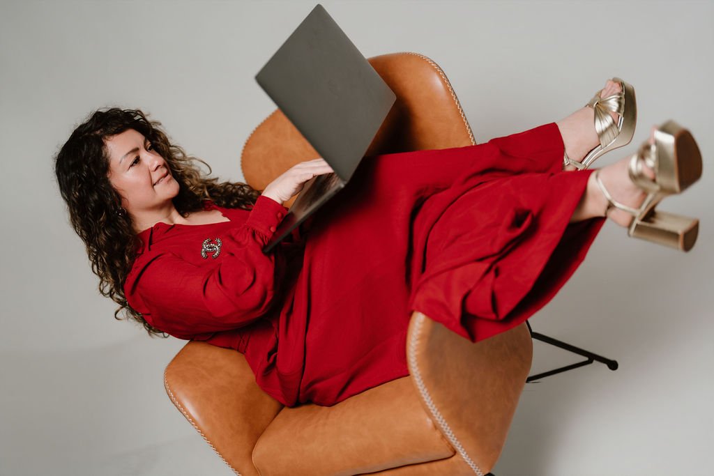 Cynthia Platero, Executive Coach in a red dress and high heels relaxing on a brown leather chair while using a laptop.