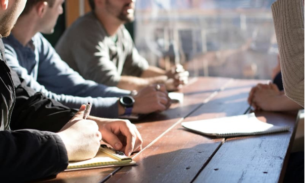 People sitting at a wooden table during a meeting or discussion, taking notes, with sunlight streaming in from a window.
