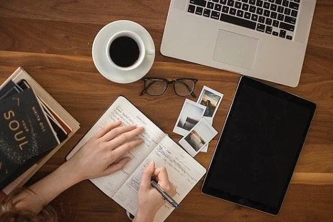 A desk with a laptop, a tablet, a pair of eyeglasses, a cup of coffee, a notebook, a pen, a book, and some printed photos, with someone writing in the notebook.