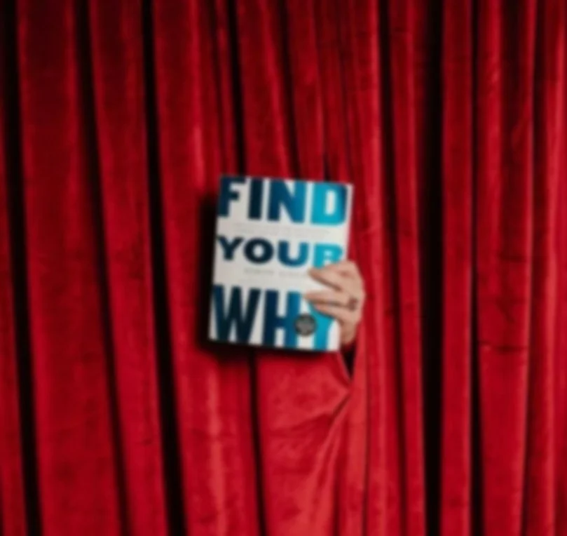 Person holding a book titled 'Find Your Why' in front of red velvet curtains.