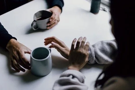Two people holding coffee mugs across a table with a person's arm reaching toward the mugs.