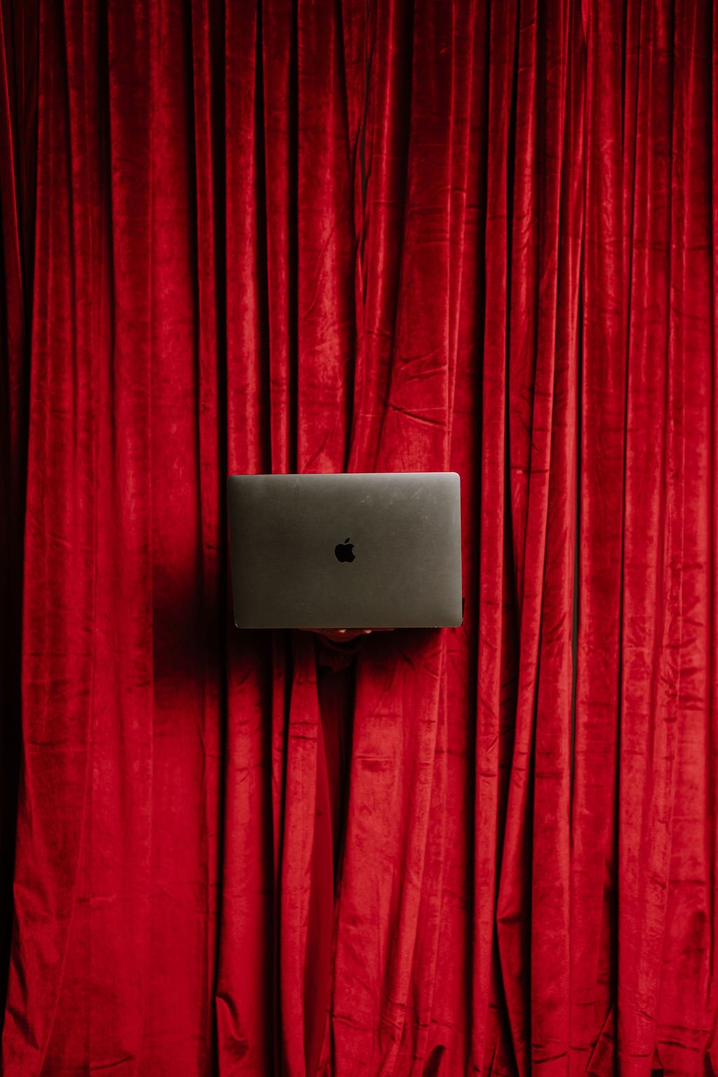 A closed MacBook laptop held in front of red stage curtains.
