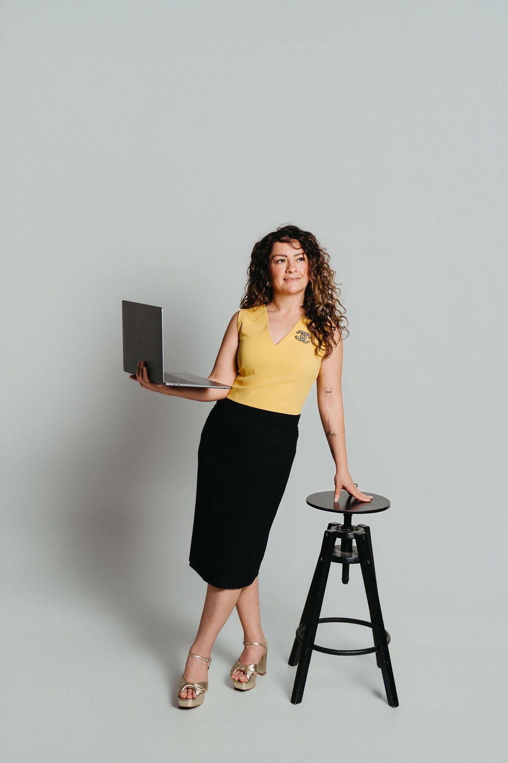 Woman wearing a yellow top and black skirt, holding a laptop in one hand and resting her other hand on a stool, standing against a plain light background.