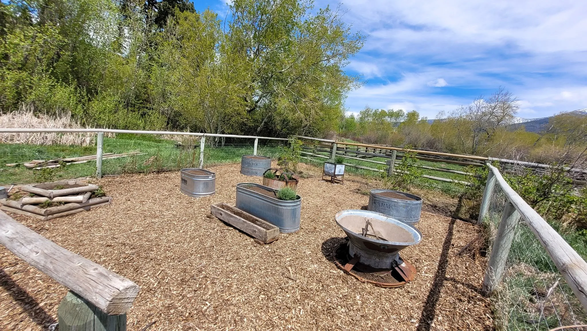 A fenced outdoor area with raised beds and metal stock tanks, surrounded by trees and mountains in the distance under a partly cloudy sky.