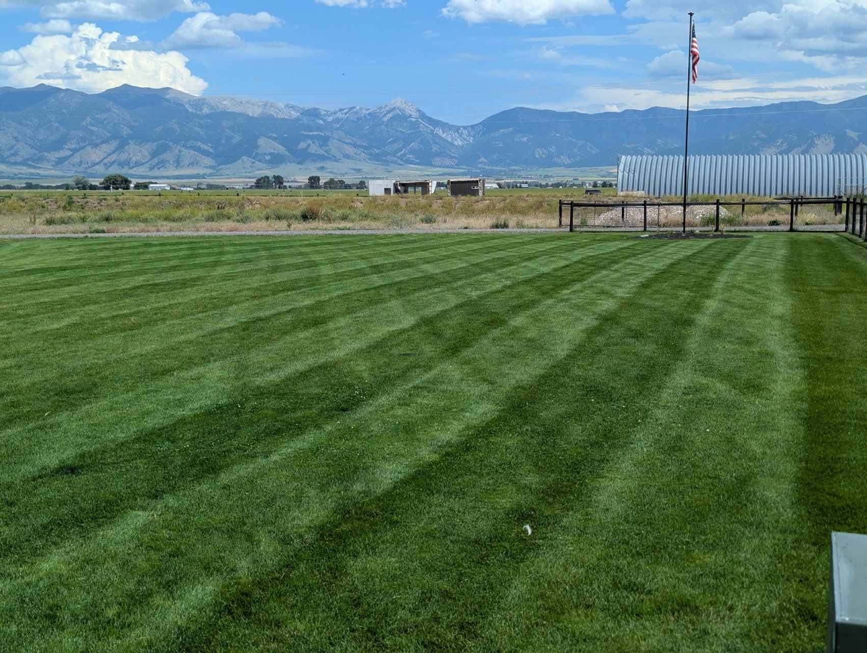 A scenic view of a well-maintained green lawn with mountain range in the background, an American flag on a tall pole, some farm structures, and a clear sky with clouds.