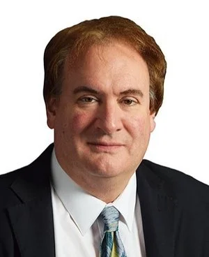 A portrait of a middle-aged man with light brown hair, wearing a black suit, white shirt, and a patterned tie, against a white background.