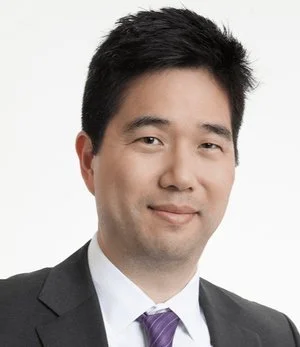 Portrait of an Asian man in a business suit with a white shirt and purple tie, smiling against a plain gray background.