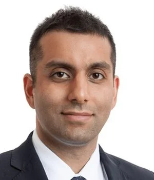 A professional headshot of a man with short dark hair, wearing a suit and tie, smiling calmly against a plain white background.