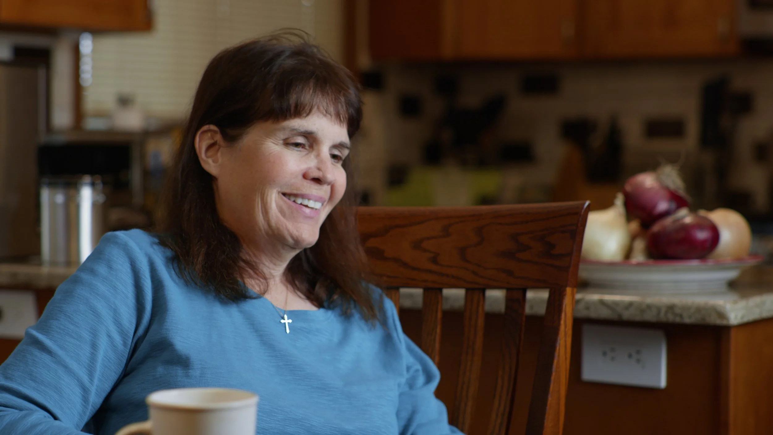A woman with dark brown hair and a blue shirt is sitting at a wooden table, smiling and looking slightly down. There is a white mug in front of her.