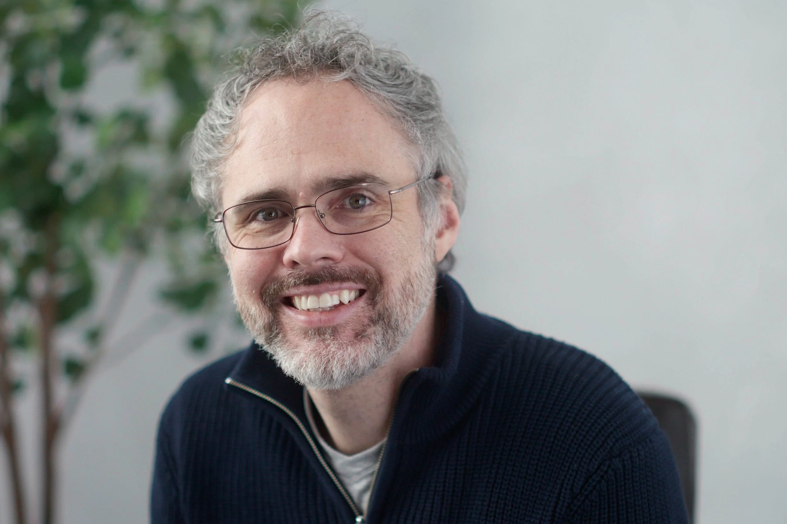 Smiling man with gray, curly hair and glasses, wearing a dark sweater, sitting in a bright room with a plant in the background.