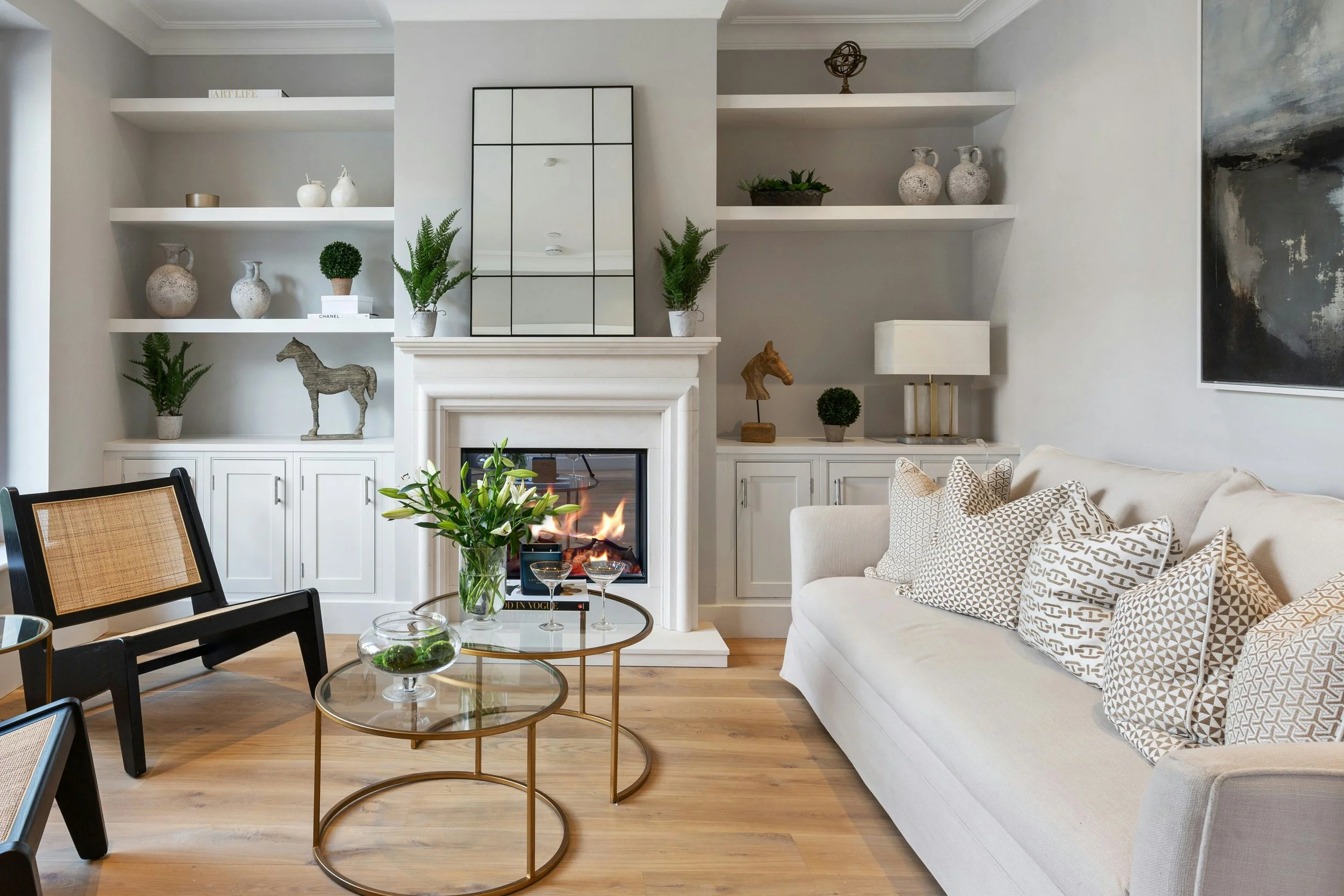 Modern living room with a fireplace, white built-in shelves with decorative vases and plants, a beige sofa with patterned cushions, black armchair, and glass-top coffee tables with a flower arrangement